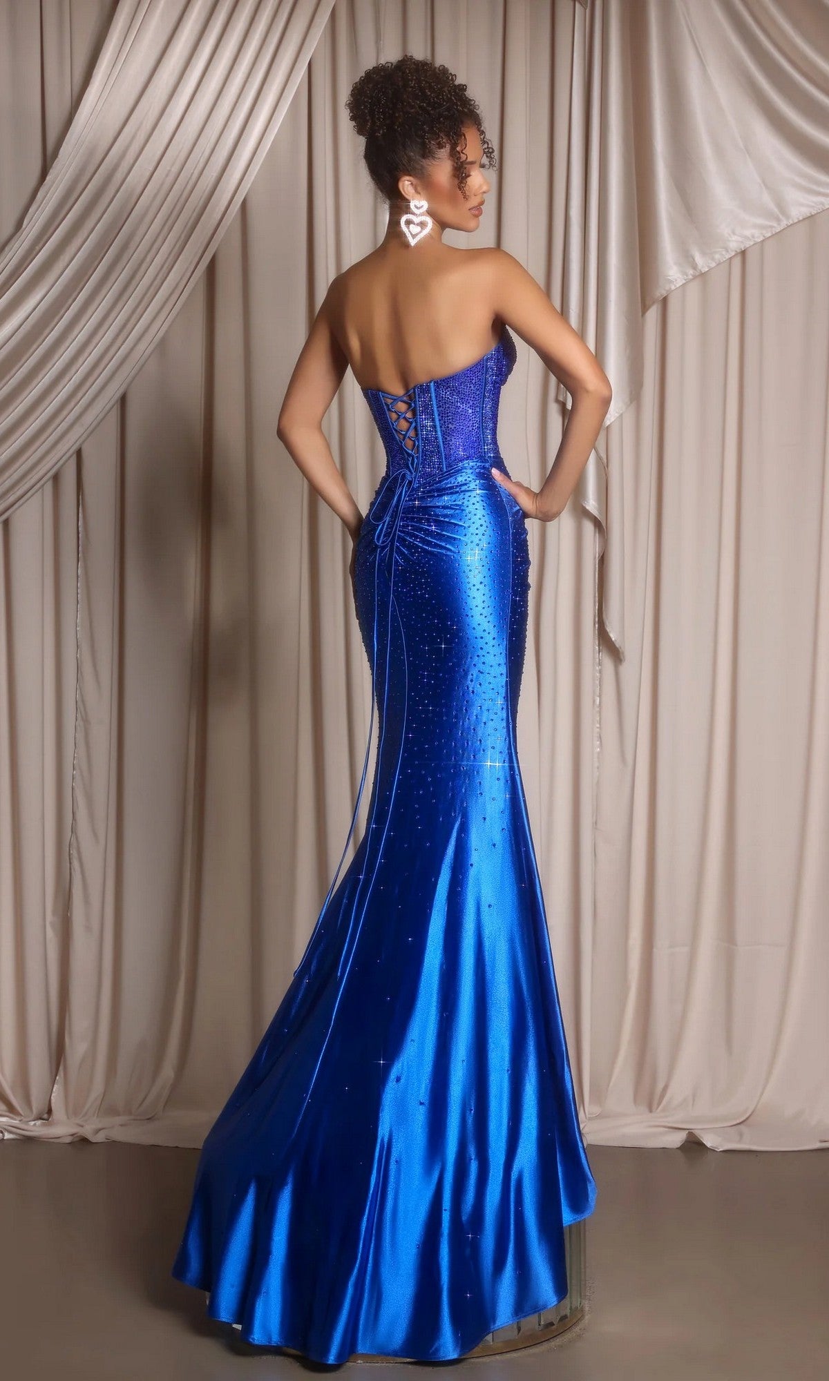 A woman stands with her back to the camera in front of beige curtains, wearing the Portia and Scarlett XO098 long satin prom dress in royal blue, which features a strapless design, lace-up back, and floor-length mermaid skirt.